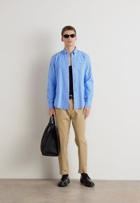 Lichtblauw overhemd, zwarte tanktop, beige broek, zwarte schoenen, een gestructureerde zwarte tas vasthoudend, met donkere zonnebril.