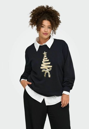 Femme aux cheveux bouclés portant un pull noir avec un motif d'arbre de Noël doré par-dessus une chemise blanche à col et un pantalon noir.