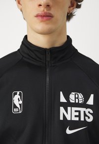 Giacca nera con zip con loghi NBA e Brooklyn Nets in bianco. Caratterizzata da un alto collare e una texture liscia e opaca. Include un dettaglio con il logo Nike.