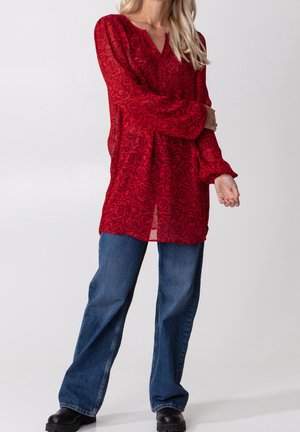 Nainen, joka pukeutunut punaiseen pitkähihainen puseroon, jossa on ruusukuvioita, sinisiin farkkuihin ja mustiin kenkiin, seisoo yksiväristä vaaleaa taustaa vasten.