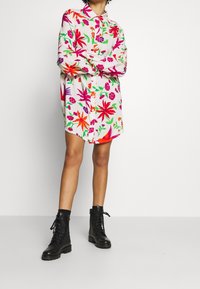 Květinové šaty košilového střihu v živých barvách s bílým základem, s zapínáním na knoflíky vpředu, dlouhými rukávy a volnějším střihem, doplněné černými vojenskými botami.