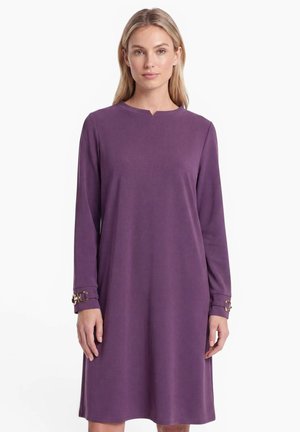 Woman wearing a long-sleeve, knee-length purple dress with gold buckle accents on the cuffs, standing against a plain white background.