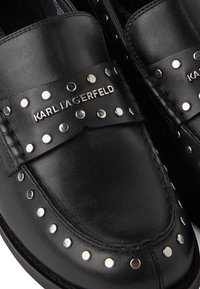 Crne kožne mokasine sa srebrnim zakovicama duž ivice i dekorativnom trakom koja sadrži ime branda u metalik slovima.