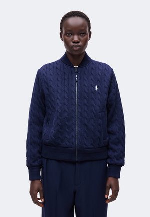 Femme portant un pull zippé en maille torsadée bleu marine avec un petit logo blanc sur la poitrine gauche, debout devant un fond uni.