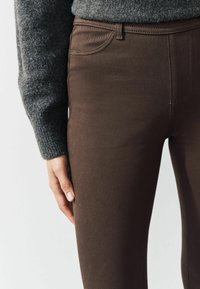 Jeans attillati marroni scuri con una superficie liscia, dotati di tasche standard e cuciture visibili lungo le cuciture. Mano appoggiata sul fianco.