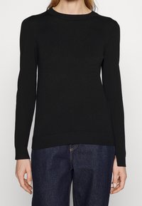 Femme portant un pull noir ajusté à manches longues et un jean en denim bleu foncé, se tenant devant un fond uni.