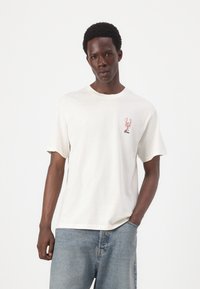 Tricou cu maneci scurte de culoare crem, cu un croi lejer și un print cu un homar roșu pe pieptul stâng. Asortat cu blugi din denim albastru deschis.