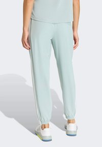 Pantalon de survêtement bleu clair avec des rayures blanches sur les côtés, des poignets élastiques et une texture douce. Associé à des chaussures de sport blanches avec des semelles bleu et vert.
