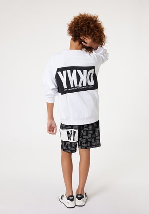 Sweatshirt blanc avec un logo noir surdimensionné au dos, associé à un short noir présentant un motif graphique blanc répétitif et des baskets blanches.