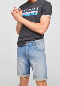 Miestenské čierne tričko s grafikou "MIAMI VICE" v ružovej a modrej farbe, kombinované s svetlo modrými džínsovými šortkami s obrúbenými manžetami a výraznými prešívacími detailmi.