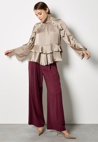 Blusa beige a balze con maniche lunghe, colletto alto e design a strati, abbinata a pantaloni larghi bordò e tacchi nudi eleganti.