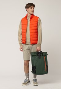 Chaleco acolchado naranja, camisa beige, pantalones cortos claros, sosteniendo una mochila verde oscuro con detalles en negro y un zipper naranja. Zapatillas con detalles en azul.