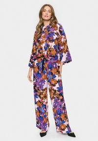 Bluzica s cvetnim printom in široke hlače v vijolični, roza, modri in rjavi barvi. Ima zadrgo na sprednji strani, kravato v pasu in široke rokave.