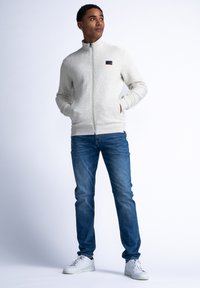 Ljusgrå zip-up jacka med en ribbad textur, två sidofickor och en liten svart logotyp. I kombination med blå jeans och vita sneakers.