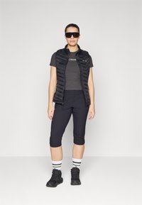 Colete puffer preto, t-shirt cinza, calças capri pretas, meias listradas e botas de caminhada pretas. Óculos de sol com armações grandes.