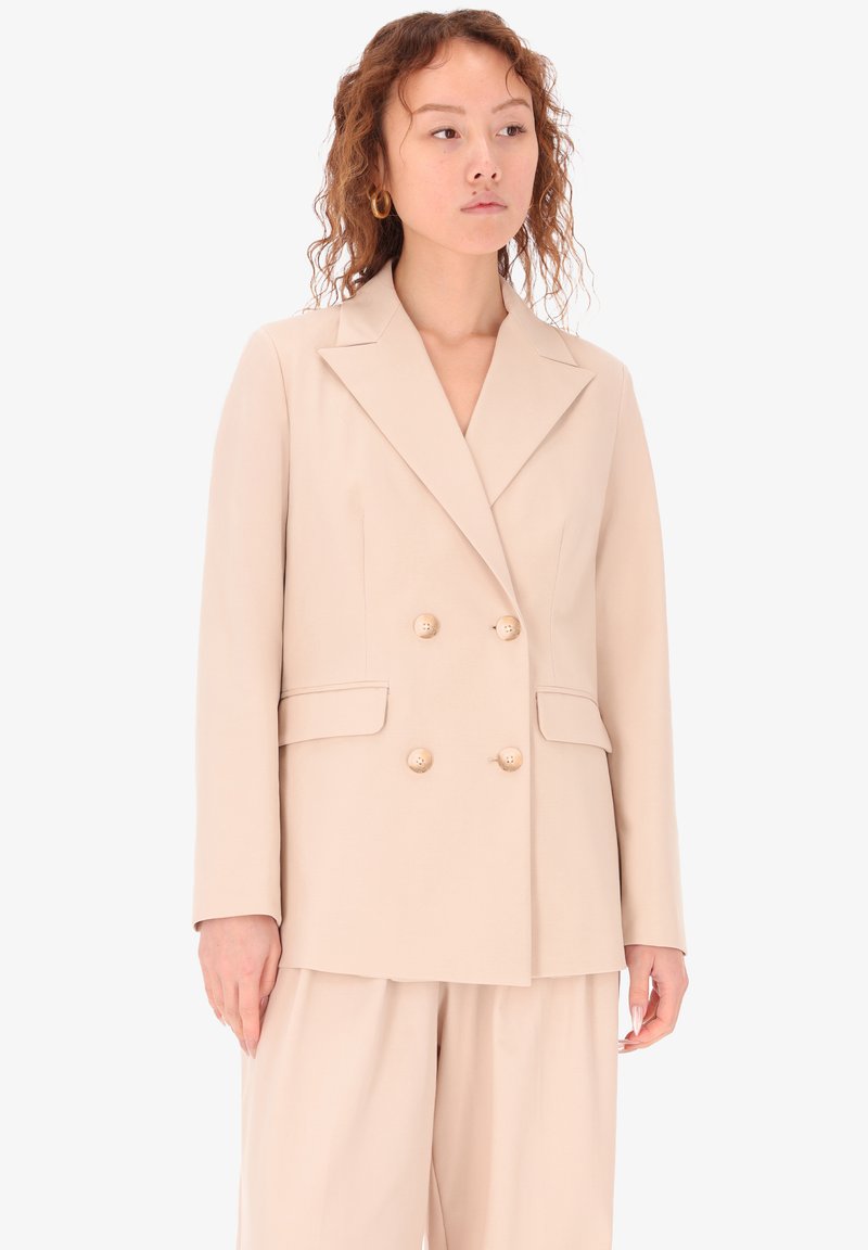 Blazer beige doppiopetto con grandi bottoni rotondi, rever a punta e due tasche frontali. Tessuto liscio con vestibilità su misura.