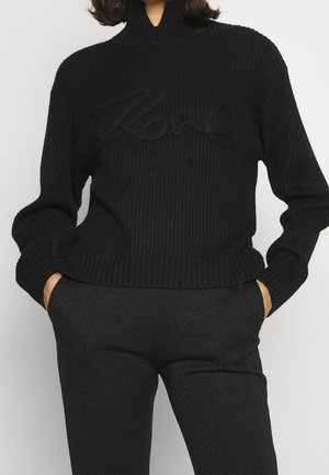 Personne portant un pull noir côtelé avec le mot « Love » brodé sur le devant et un pantalon noir, les mains dans les poches, sur un fond uni.