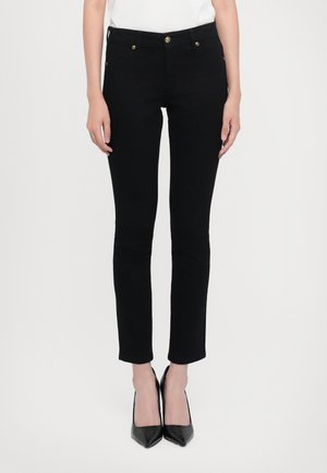 Mujer con pantalones negros ajustados combinados con tacones altos negros de punta afilada y una blusa blanca, posando frente a un fondo liso.