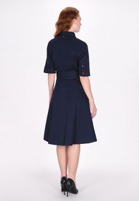 Rochie cămașă de culoare albastru marin, cu mâneci scurte, talie ajustată și fustă evazată; prezintă detalii cu nasturi și o textură netedă, țesută.