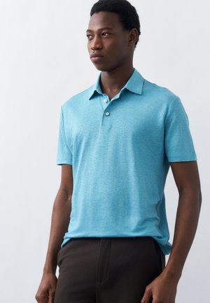 Άνδρας που φοράει ανοιχτό μπλε κοντομάνικο πόλο πουκάμισο και σκούρο παντελόνι, κοιτάζοντας προς τα πλάγια μπροστά σε απλό λευκό φόντο.