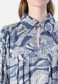 Camicia blu a fantasia con colletto, apertura a chiave e design plissettato con motivi floreali. Dettagli arricciati lungo le maniche.