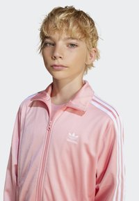 Rosa treningsjakke med hvite striper, glidelås og en liten logo. Myk tekstur og avslappet passform, passer for hverdagsbruk.