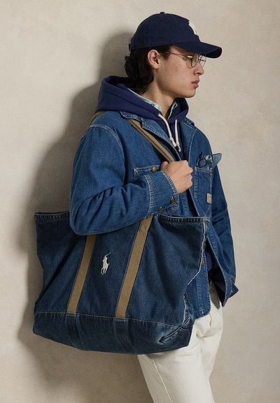 Νεαρό άτομο με τζιν μπουφάν, σκούρο μπλε φούτερ με κουκούλα, καπέλο και γυαλιά, κρατώντας μεγάλη τσάντα tote από τζιν με μπεζ λουριά και λευκό λογότυπο.