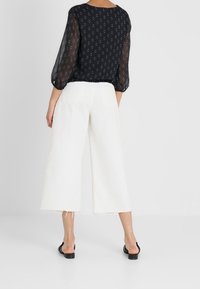 Svart genomskinlig blus med diamantmönster och puffärmar, matchad med vita vida jeans med fransar vid fållen. Skorna är svarta sandaler.