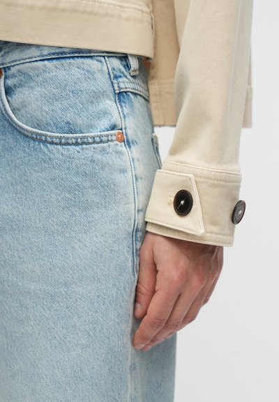 Χέρι ακουμπισμένο δίπλα στην τσέπη από τζιν τζινεζάκι ανοιχτού γαλάζιου χρώματος, φοράει μπεζ μανίκι μπουφάν με δύο σκοτεινά κουμπιά στο μανίκι.