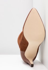 Zapato de tacón alto de ante marrón con punta afilada y un acento de cuero suave. La suela es de un tono claro de beige con un patrón de agarre texturizado.
