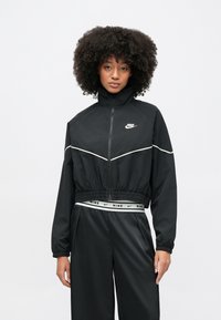 Μαύρο μπουφάν Nike με ψηλό γιακά, φερμουάρ μπροστά, ελαστική μέση και λευκές γραμμές διακόσμησης. Συνδυασμένο με μαύρο, χαλαρό παντελόνι.