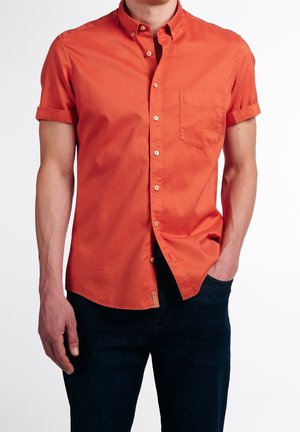 Mann trägt eng anliegendes oranges Kurzarmhemd mit Brusttasche, hochgekrempelten Ärmeln und dunklen Jeans, steht mit einer Hand in der Tasche.