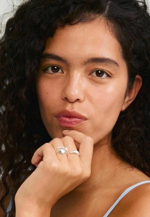 Silver rings featuring a smooth, teardrop shape and a shell-like design on the fingers, with curly, dark hair framing the face.