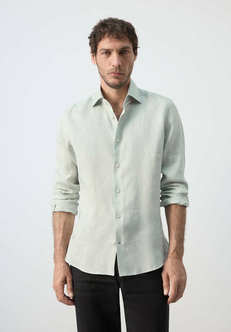 Homme aux cheveux courts et bouclés, portant une chemise vert clair à manches longues avec les manches retroussées, debout devant un fond clair uni.