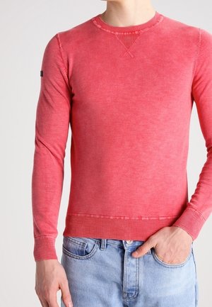 Hombre con un suéter rojo ajustado de manga larga y vaqueros azul claro, con una mano en el bolsillo.