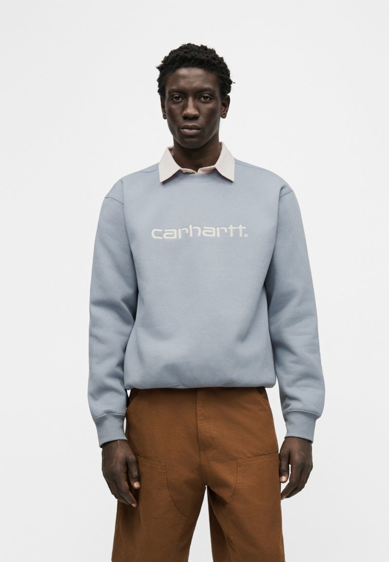 Mann steht und trägt ein hellblaues Carhartt-Sweatshirt über einem beigen Hemd mit Kragen und braune Hose vor weißem Hintergrund.