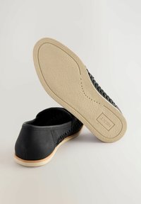 Chaussures noires en toile à enfiler avec une tige en cuir souple et une semelle en caoutchouc texturée, présentant une bordure beige et le logo de la marque sur la semelle.