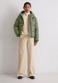 Zielona puchowa kurtka z kapturem, kremowy sweter o fakturze warkocza oraz beżowe szerokie spodnie sztruksowe. Całość dopełniają czarne buty.