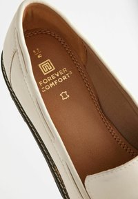Vaaleat slip-on -kengät, joissa on pyöristetty kärki. Kengissä on ruskea nahkainen pohjallinen, hienovarainen ompelu sekä teksturoitu musta ulkopohja. Kultainen brändäys näkyvissä.