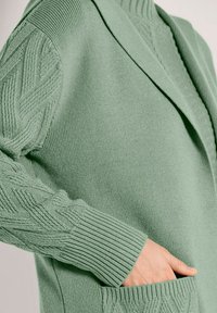 Hand rustend in de zak van een groene gebreide cardigan met kabelpatronen op de mouwen en geribde manchetten.