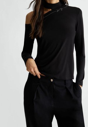 Femme portant un haut noir à manches longues avec une épaule dénudée et un pantalon noir, posant avec une main dans la poche et l'autre près de la taille.