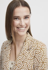 Braun gemusterter Blazer mit geometrischem weißen Design. Der Stoff hat eine glatte Textur und eine taillierte Passform, mit geschlitzten Revers.