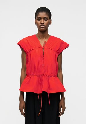 Kvinde iført en skrigrød, ærmeløs top med V-udskæring, snørebind, indsnævret talje og peplumkant, kombineret med sorte bukser.