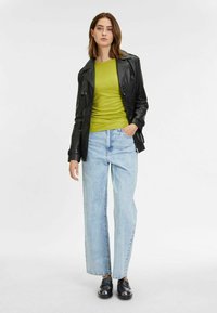 Giacca di pelle nera, top a costine verde brillante, jeans a gamba larga blu chiaro e mocassini neri; modello in piedi contro uno sfondo bianco semplice.