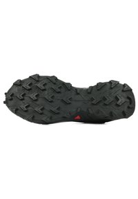 Black rubber shoe sole featuring deep, multidirectional treads, a textured surface, and a red triangular accent near the heel.