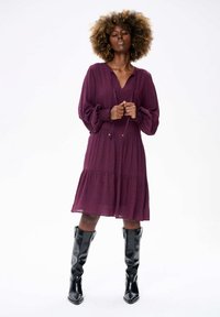 Bordeaux jurk met ballonmouwen, V-hals en knoopdetails bij de taille, gelaagd met een uitlopende rok; gecombineerd met zwarte kniehoge laarzen.