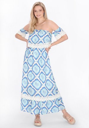 Fiatal nő áll, kezét a csípőjén tartva, kék-fehér mintás váll nélküli maxi ruhát és ékdetálos szandált visel.