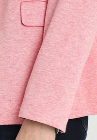 Tessuto rosa con una texture e un delicato motivo mélange. La manica presenta un polsino ripiegato e una piccola tasca a toppa. Il materiale sembra morbido.