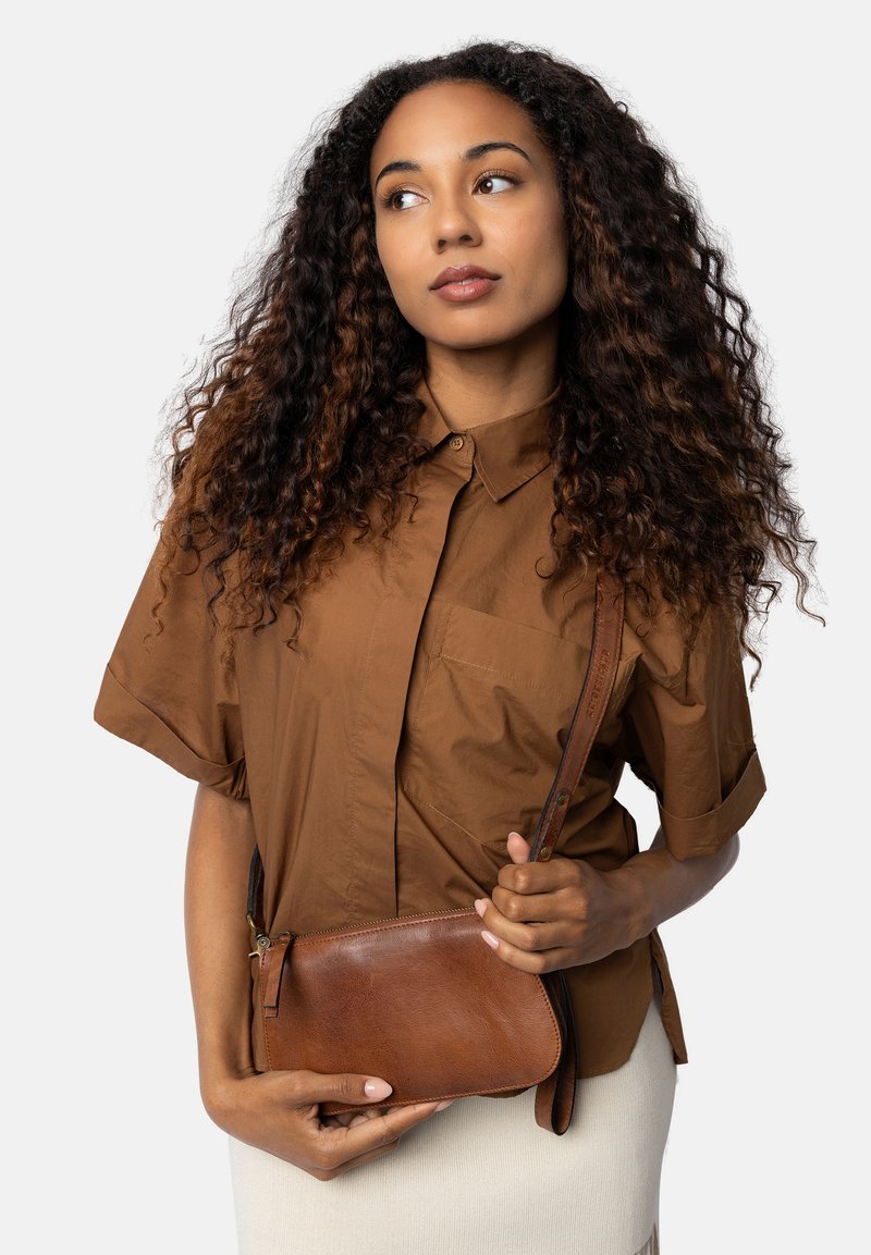 Sac en cuir marron en bandoulière avec une fermeture éclair, design minimaliste et sangle ajustable, tenu contre une tenue de couleur claire.