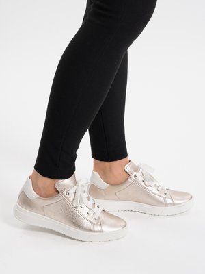 Person trägt metallisch roségoldene Turnschuhe mit weißen Sohlen und Schnürsenkeln, kombiniert mit schwarzen, eng anliegenden Hosen vor einem schlichten Hintergrund.